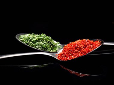 Kırmızı biber tozu ve siyah bir yüzeye yansıyan kaşıklardaki maydanoz tanelerinin yakın çekim fotoğrafı..