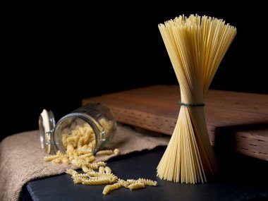 Kurutulmuş spagetti eriştelerinin ayakta durduğu ve cam kavanozdan sarmal şehriyelerin döküldüğü bir fotoğraf..