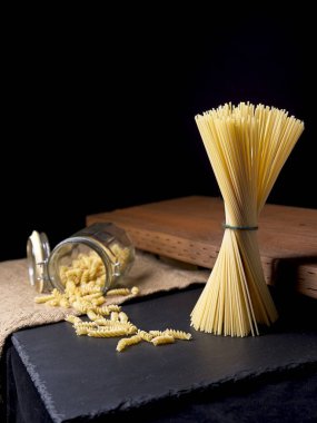 Kurutulmuş spagetti eriştelerinin ayakta durduğu ve cam kavanozdan sarmal şehriyelerin döküldüğü bir fotoğraf..