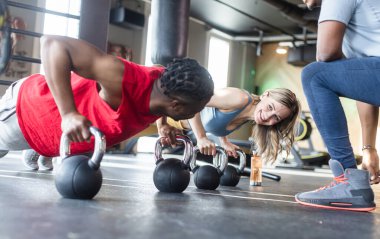 Modern spor salonunda kadın ve erkek, koçun gözetiminde kettle çanlarıyla şınav çekiyorlar.