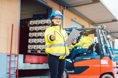 Endüstriyel kıyafetli bir kadın bir forkliftin önünde duruyor ve malları bir depodan taşıyor.