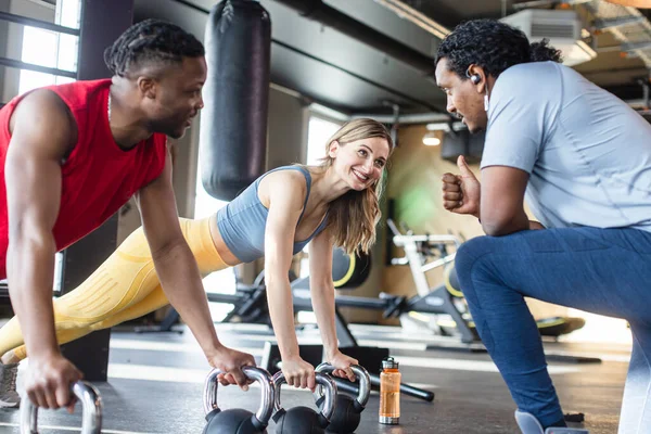 Diversity woman and man in modern gym doing push-ups with kettle bells under supervision of coach