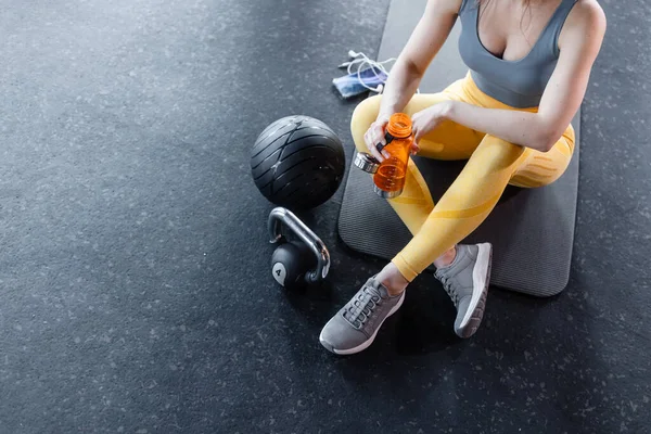 一位穿着运动服的健康漂亮的年轻女子坐在体育馆的地板上 旁边的水壶铃边 手里拿着电话和水瓶 图库图片