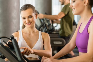 Biri koşu bandında koşan, diğeri de spor aletinin dokunmatik ekranına parametreler giren çok formda ve güzel iki genç kadın.