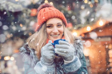 Kış pazarında Noel ağacının önündeki kadın kameraya bakarak sıcak şarap içiyor.