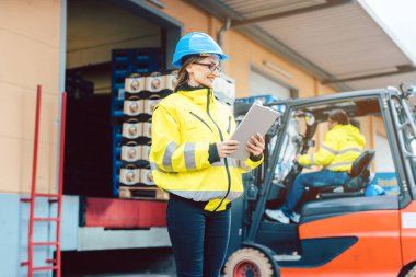 Endüstriyel kıyafetli bir kadın bir forkliftin önünde duruyor ve malları bir depodan taşıyor.