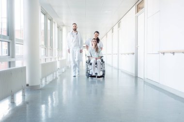 Doktor, hemşire ve tekerlekli sandalyedeki hasta hastane koridorunda yürüyor.