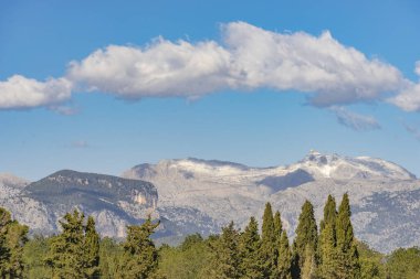Karla kaplanmış bir dağ sırasının ihtişamı yoğun bir yeşil ormanın üzerinde duruyor. Mavi gökyüzüne karşı kabarık bulutlarla süslenmiş bu panoramik sahne,