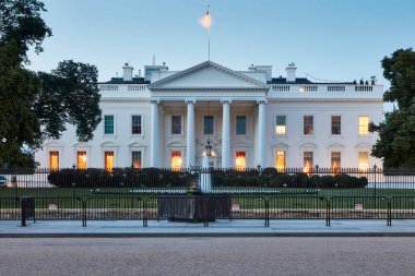 Washington, D.C. 'deki Beyaz Saray' ın net ve simetrik bir görüntüsü, şafakta çekildi. Yumuşak sabah ışığı, bu tarihi binanın pencerelerinden yayılan sıcak ışıkla çelişiyor. ABD bayrağı tepede dalgalanıyor..