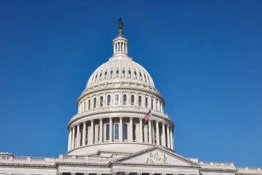 Washington, D.C. 'deki Birleşik Devletler Kongre Binası' nın sembolik kubbesi. Amerikan bayrağı kubbenin önünde dalgalanıyor. Parlak, berrak bir mavi gökyüzüne yakalanmış.