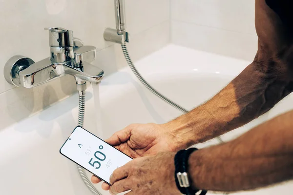 スマートホーム概念 男は彼の携帯電話上のアプリを使用して温水温度を制御します ロイヤリティフリーのストック写真