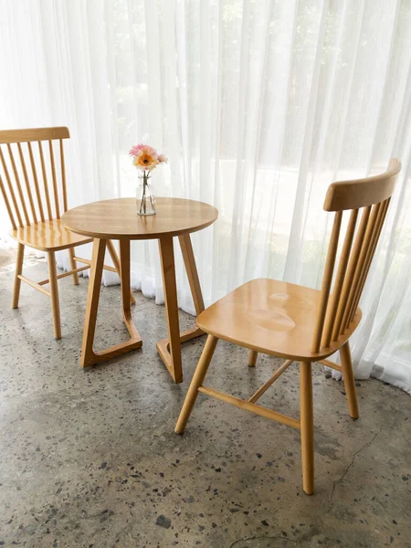 The wooden chair and table set in the minimal style with the blooming flower near a window decorated by the white curtain in the small coffee shop, front view for the copy space.