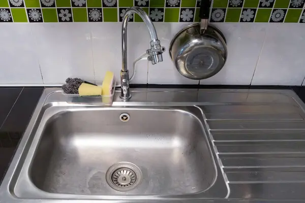The clean metal sink with the chrome tap after washing on the modern counter, stainless steel pot and sponges and equipment for washing near the sink, inside the kitchen of the urban house, front view for the copy space.