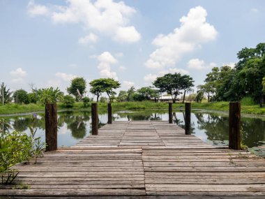 Kırsal bir ahşap iskele, bereketli bir bahçedeki sakin bir gölün üzerine uzanır, ağaçları ve gökyüzünü yansıtır, doğa severler ve rahatlama arayanlar için huzurlu bir dinlenme yeri sağlar..