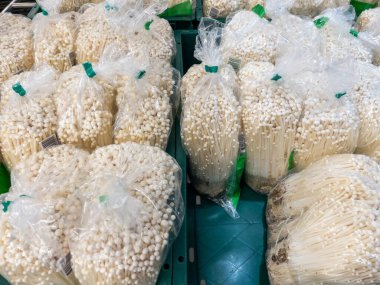 Clusters of white enoki mushrooms packed in transparent plastic bags, secured with green ties, and arranged on green crates for sale in a supermarket. Ideal for healthy meals, showcasing fresh produce abundance.