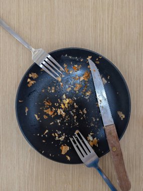 A black ceramic plate with crumbs left from the bakery, accompanied by a fork and knife, placed on a wooden table after a finished meal.