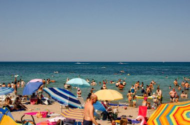 Bilinmeyen müşterileri ve şemsiyelerini gösteren kumsalın fotoğrafı ve güneşli bir havada ufuk çizgisi.