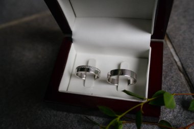 İki gümüş alyans. Zirkonlu kadınlar ve erkeksiz erkekler. Beyaz satenle kaplı ahşap bir kutuda. Gri arkaplanda ve çevresinde bulanık bitkiler var..