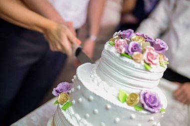 Üç katlı düğün pastasını altın kurdele, fiyonk ve dekoratif çiçeklerle kesiyor ve boncuklu beyaz bir tabanı var.