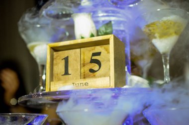 15 Haziran tarihi ahşap kutunun üzerine siyah yazılıyken, arka planda dumanın çıktığı cam bardaklar var.