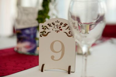 Kırmızı masa örtüsü ve kırmızı peçetelerle süslenmiş bir yemek masası, gri bir şamdan, üç gül ve ahşap bir standa oyulmuş dokuz numara.