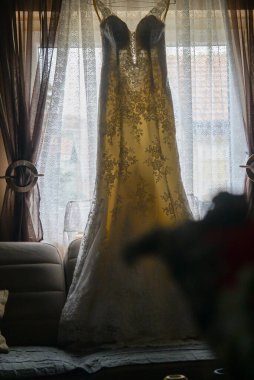 Dar, beyaz bir gelinlik askıda, pencerenin önünde güçlü bir tezgah lambasıyla çekilmiş.