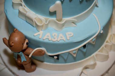 Mavi yuvarlak, üzerinde oyuncak ayılar olan iki katlı doğum günü pastası, üstünde bir numara ve erkek adı Lazar.