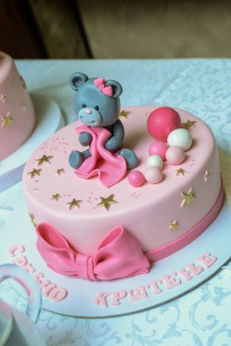 Yuvarlak pembe bir doğum günü pastası pembe ve beyaz süslemelerle balonları çekiyor ve üstünde pembe bir battaniye tutan bir oyuncak ayı..