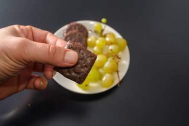 Beyaz üzümler ve çikolatalı bisküviler, aynı tabakta iki farklı sağlıklı ve sağlıksız şeker ve koyu arkaplandaki sözde yiyeceklerle aynı.