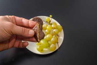 Beyaz üzümler ve çikolatalı bisküviler, aynı tabakta iki farklı sağlıklı ve sağlıksız şeker ve koyu arkaplandaki sözde yiyeceklerle aynı.