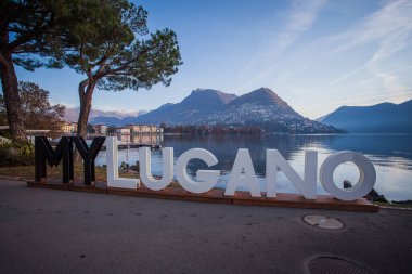 Lugano, İsviçre - 12 Kasım 2021: Lugano Gölü kıyılarında sonbahar panoraması. Önplanda şehrin adı yazılı metal bir yazı var.