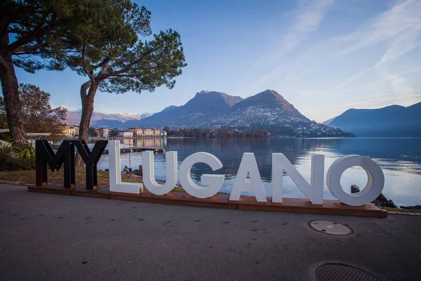 Lugano, İsviçre - 12 Kasım 2021: Lugano Gölü kıyılarında sonbahar panoraması. Önplanda şehrin adı yazılı metal bir yazı var.