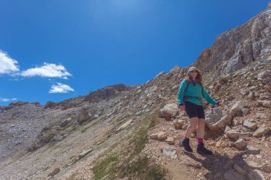 Güneş gözlüklü, uzun hava ve şapkalı genç bir kadın dolomit tepelerinin ortasında yürüyor, Latemar, Trentino, İtalya. Gezici fotoğrafçılık ve açık hava sporları konsepti