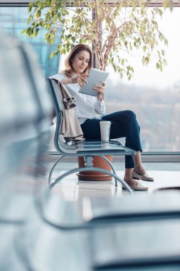 Seyahat eden genç bir kadın tablet kullanıyor ve havaalanının kalkış salonunda bekliyor..