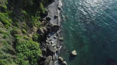 Geniş kayalar, yeşil yapraklar ve berrak mavi deniz suyuyla engebeli bir sahil şeridi perspektifi..