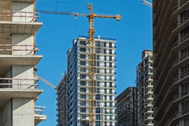 Gelişiminin çeşitli aşamalarında çok katlı binaların yer aldığı inşaat sahasında, açık mavi gökyüzüne karşı yapılar üzerinde vinçler yükseliyor..