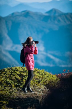 Sırt çantalı kadın yürüyüşçü güneşli bir günde DSLR kamera kullanarak manzaralı mavi tepeleri yakalıyor..