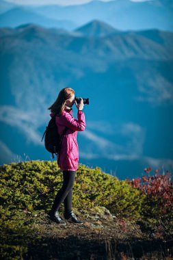 Sırt çantalı kadın yürüyüşçü güneşli bir günde DSLR kamera kullanarak manzaralı mavi tepeleri yakalıyor..