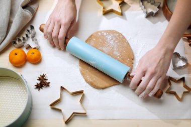 Person uses a blue rolling pin to flatten gingerbread dough surrounded by Christmas cutters and spices.