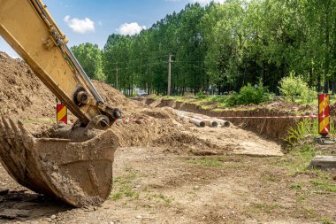 İnşaat alanının yanında bir kazıcı kovası, siperdeki yeni fiber cam su boru hattı. Eski paslı boruların yenisiyle değiştirilmesi. Rüzgarda sallanan kırmızı beyaz uyarı şeridiyle çevrili.