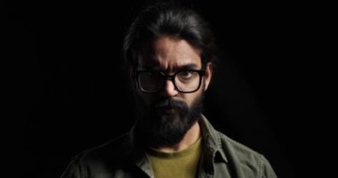 Gözlüklü ve sakallı kızgın bir gencin siyah arka planda izole edilmiş kameraya ciddi ciddi baktığı yavaş çekim.