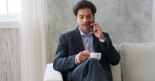 若いビジネスマンがスマートフォンで話し ソファでコーヒーを飲む — ストック動画