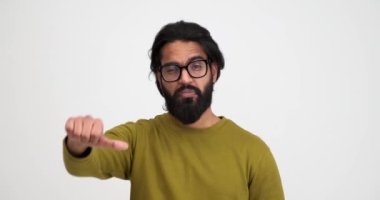 Gözlük takan üzgün sakallı bir gencin portresi beyaz arka planda tek başına dikilirken baş parmak hareketi gösteriyor.