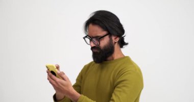 Sinirli sakallı genç adam, izole edilmiş beyaz arka plana karşı ayakta dururken hayal kırıklığı içinde cep telefonu çığlığı atmaya çalışıyor.