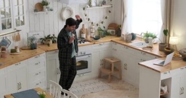 Mutfakta spatulayla dans edip şarkı söylerken dizüstü bilgisayarında müzik çalan neşeli bir adam.