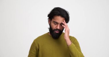 Yorgun sakallı genç adam beyaz arka planda tek başına dikilirken gözlük takıyor ve acı içinde başını ovuyor.