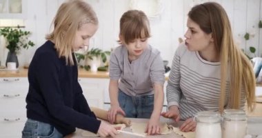 Beyaz tenli genç anne, evde mutfak masasındaki kurabiyeler için hamur kesmeyi öğretiyor ve onlara yardım ediyor.