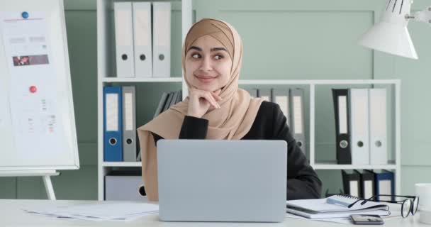 ヒジャーブのアラブ人実業家を確信し オフィスの机に座っている 広い笑顔で 彼女は積極性を示す 側面に向かって見ています 彼女はカメラに目を向け輝く笑顔を保ち — ストック動画