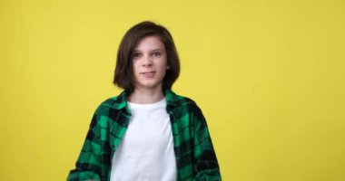 Ekose gömlek giyen genç beyaz çocuk sarı bir arka planda duruyor, öfkeyle hareketler yapıyor ve duygularını yoğun yüz ifadeleriyle ifade ediyor..