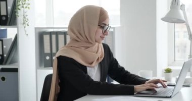 Genç Arap iş kadını tesettür takıyor, kendine güven ve pozitif enerji yayıyor. Ofis masasında oturuyor, özenle dizüstü bilgisayarına yazıyor, yukarı bakıyor, geniş bir gülümsemeyle kameraya bakıyor..